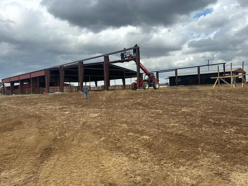 Red iron frame of an arena cover with a telehandler setting purlins, storm clouds overhead