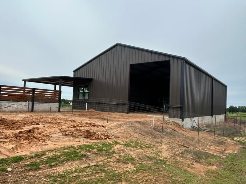 Dark brown metal barndominium shell with an open bay door, side patio and a cedar slat fence