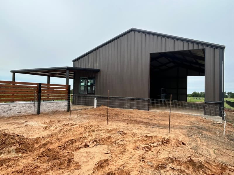 Dark brown metal barndominium shell with an open bay door and covered side patio behind a cedar fence