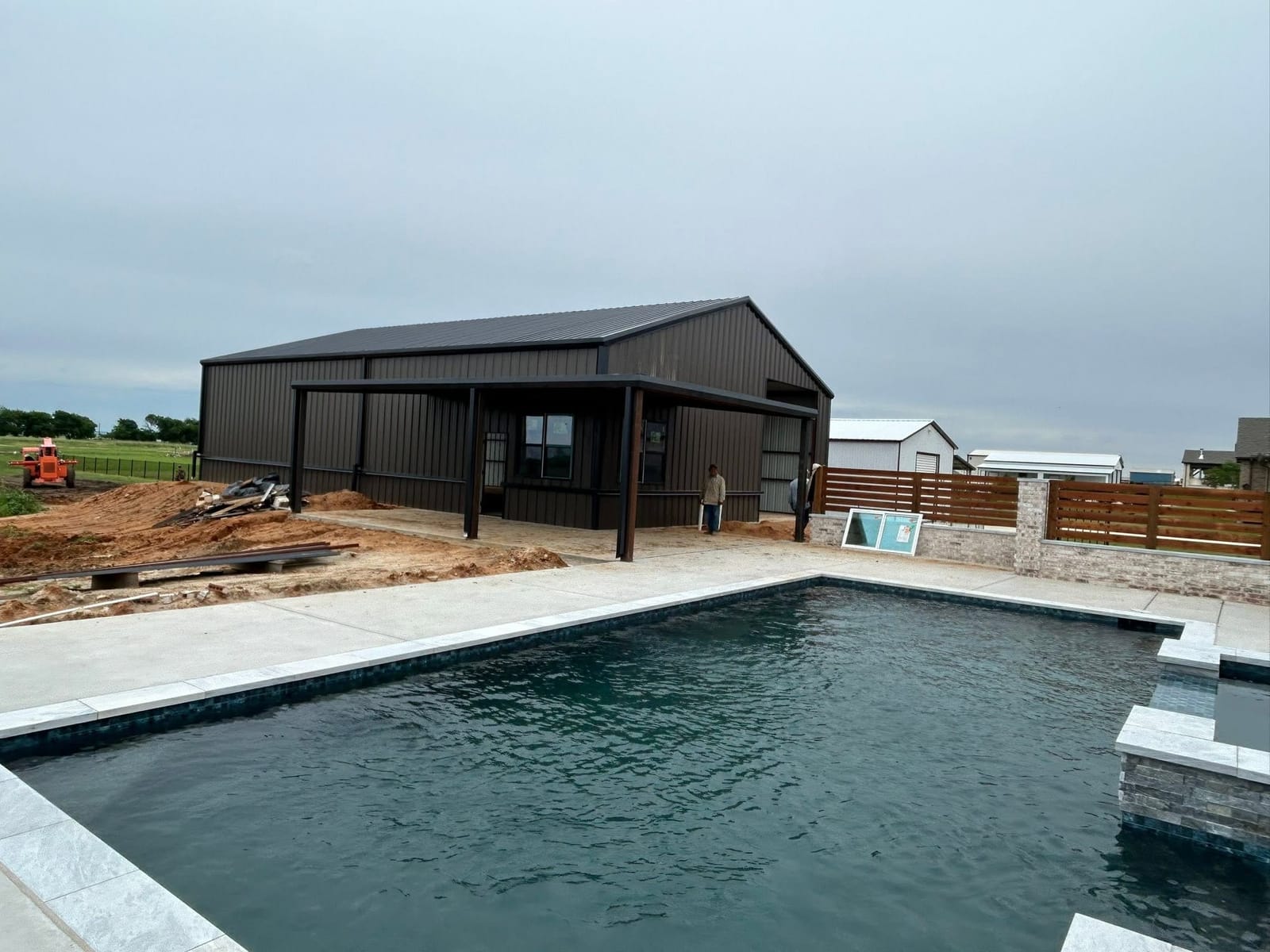 Dark brown metal barndominium behind a newly finished swimming pool and cedar fence