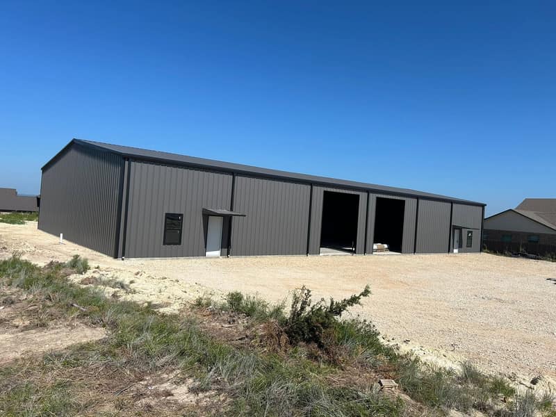 Large dark brown metal shop with two bay openings, seen from farther back across the gravel