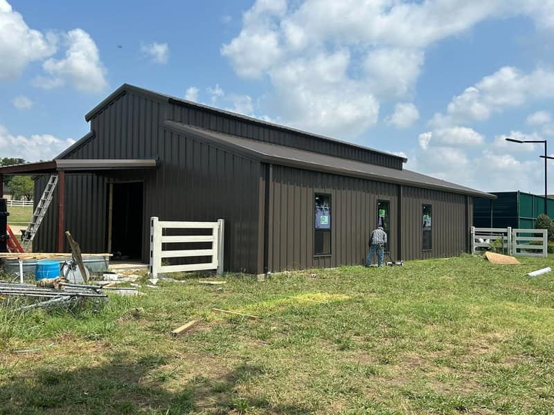 Dark brown metal shop with a covered lean-to and white fence panels on a green lawn