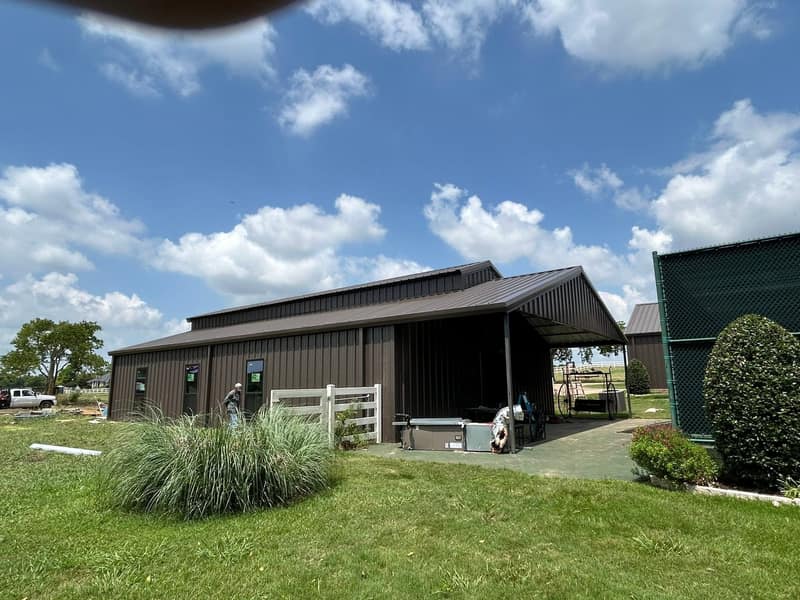 Dark brown metal shop with a covered lean-to and hot tub, seen across the lawn