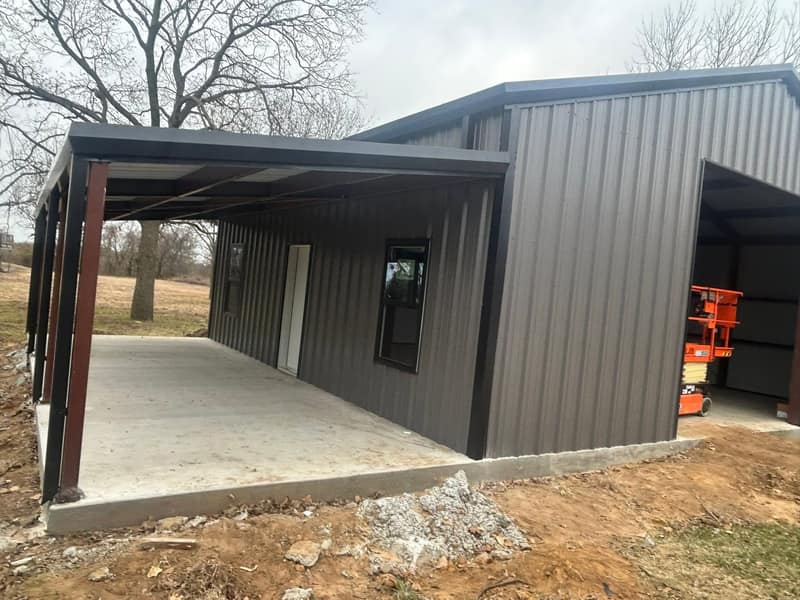 Dark brown metal shop with an open-sided lean-to on a fresh concrete slab, scissor lift inside