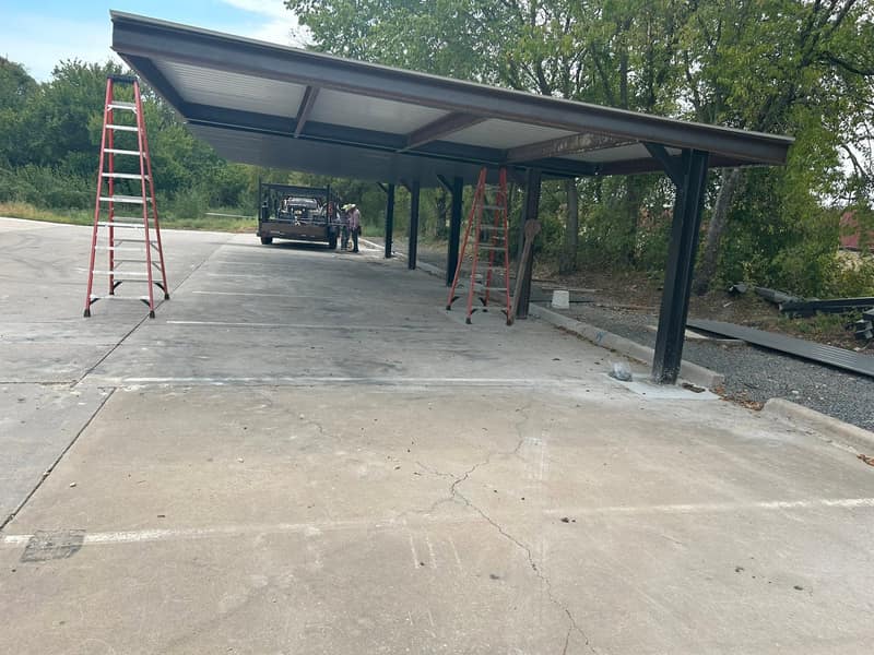 Drive-through metal carport on black posts over a concrete drive, ladders set for the trim