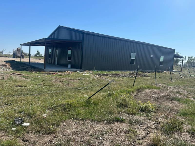 Charcoal metal barndominium with a covered porch on the end, seen across tall grass