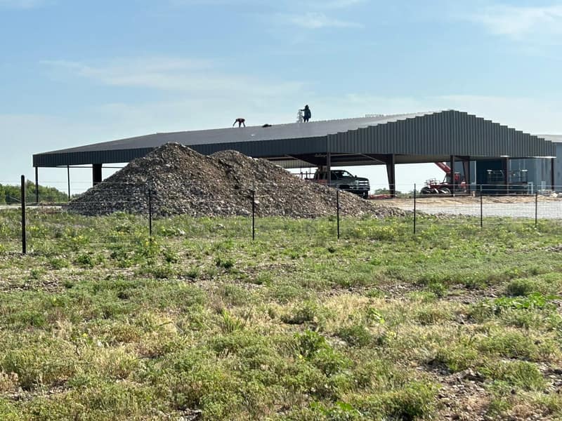 Large charcoal open-sided commercial cover with a gravel pile in front, fence line in the foreground