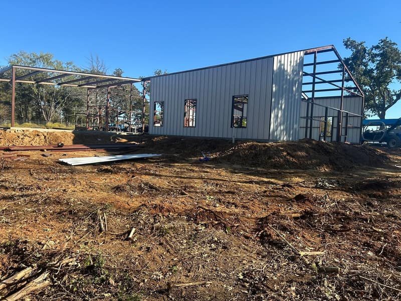 Light gray metal shop mid-build with the red iron frame still open at one end