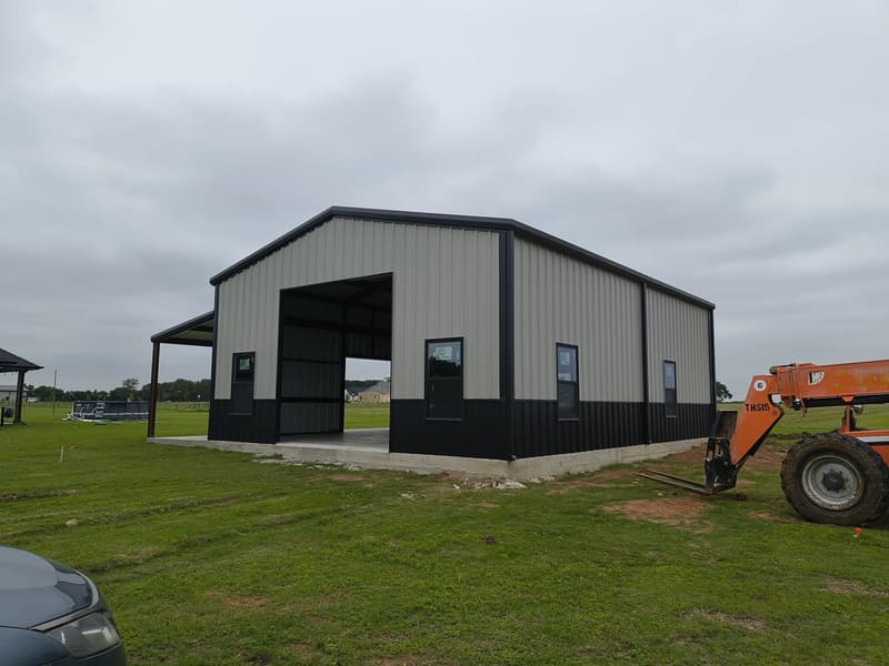 Gray and black metal shop with a tall roll-up door opening and four windows, telehandler parked alongside