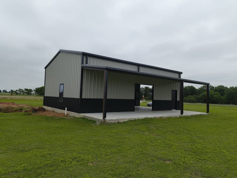Light gray metal shop with black wainscot and a covered lean-to on a fresh concrete slab