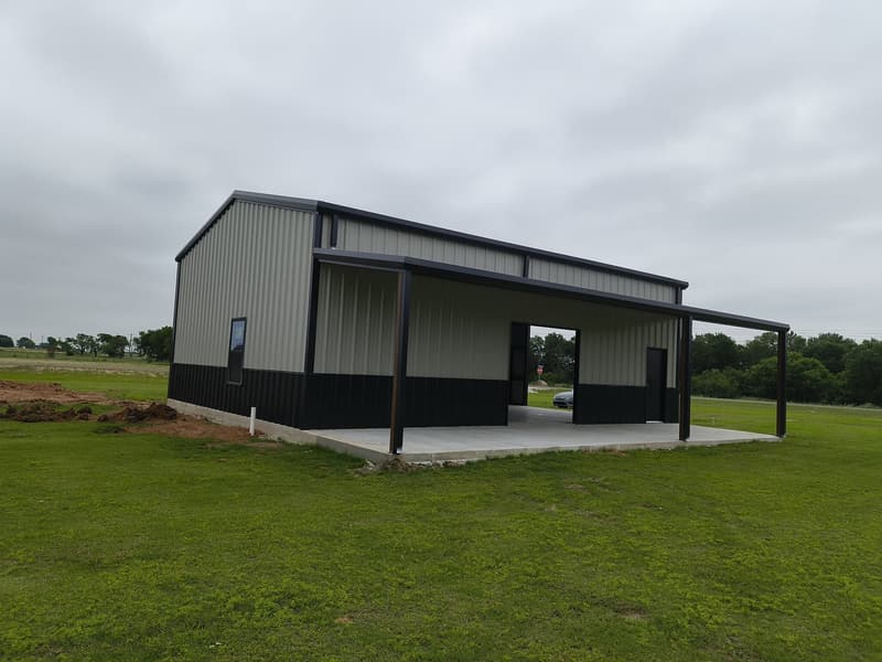 Light gray metal shop with black wainscot and a covered lean-to, viewed from the yard