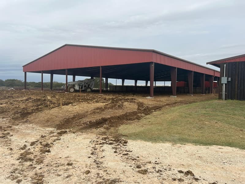 Red open-sided metal arena cover with tractor and equipment underneath, dirt freshly graded