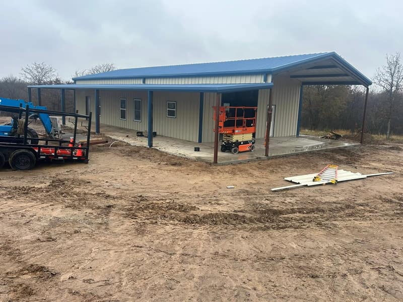 End of a tan metal building with blue trim and a covered lean-to, scissor lift and trailer alongside