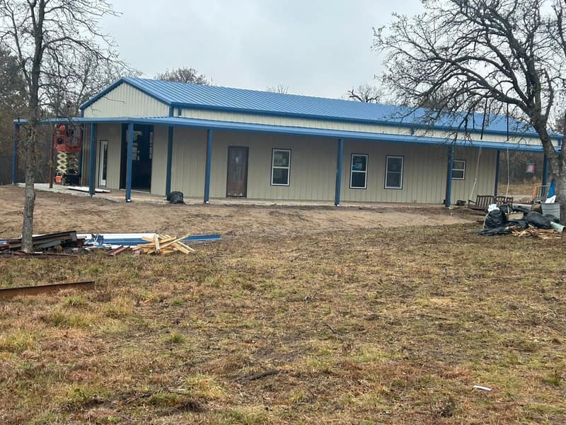 Long tan metal building with blue trim and a full-length covered porch, seen across the yard