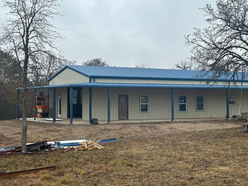 Long tan metal building with blue trim and a covered porch, shaded by bare oak trees