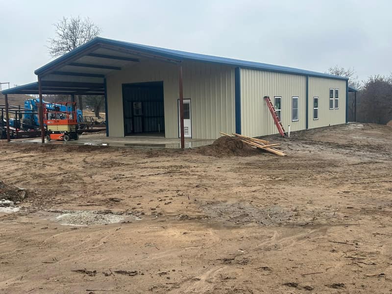 Tan metal building with blue trim, windows along the side wall and a lift working under the lean-to