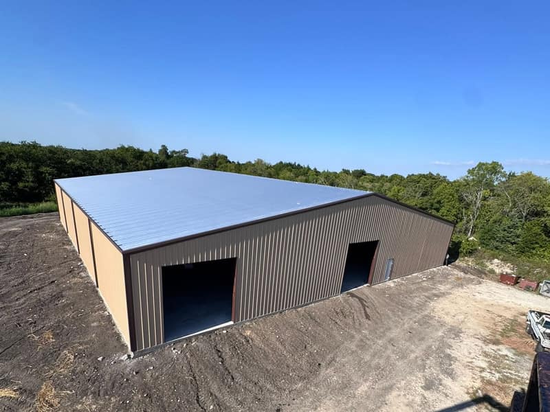Tan metal shop with a silver roof and two bay openings set into a hillside, treeline behind