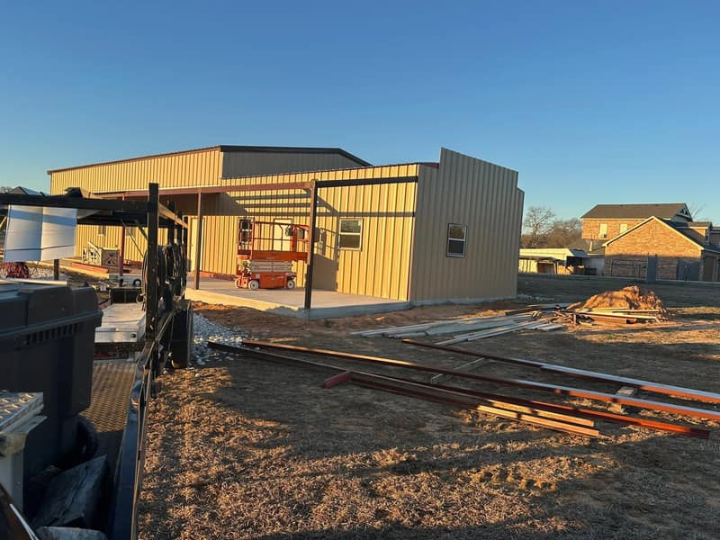 Tan metal shop with a covered lean-to in evening light, scissor lift working underneath