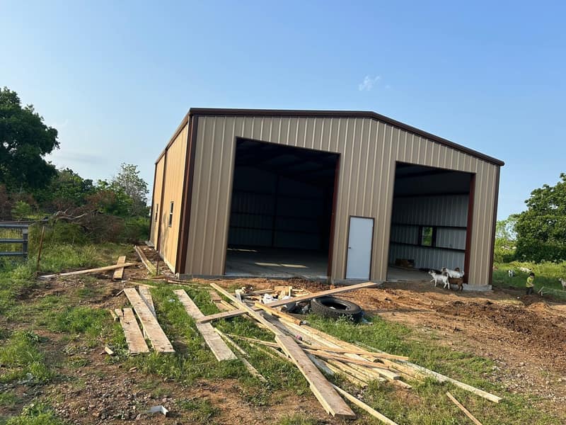 Tan metal shop with two open bays and a walk door, lumber stacked in front