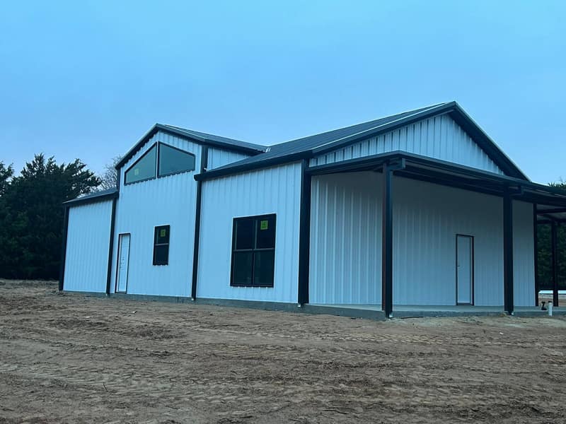 White metal barndominium with black trim, a gable window and a covered side porch on graded dirt