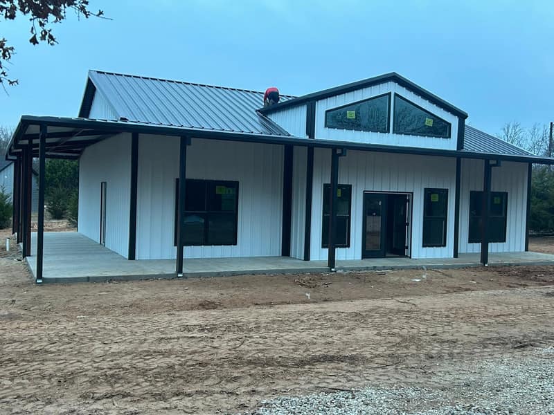White metal barndominium with a wide covered porch, black trim and double glass doors