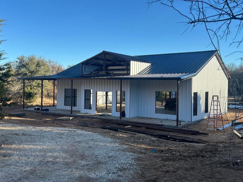White metal barndominium with a long covered porch, steel and lumber staged on the ground in front