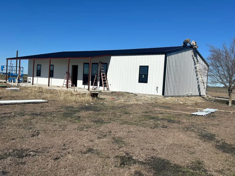 White metal barndominium with a charcoal roof and covered porch, crew working on the ridge with ladders set