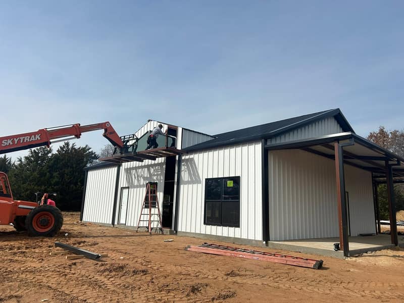 White metal barndominium under construction with a telehandler lifting panels to the roof