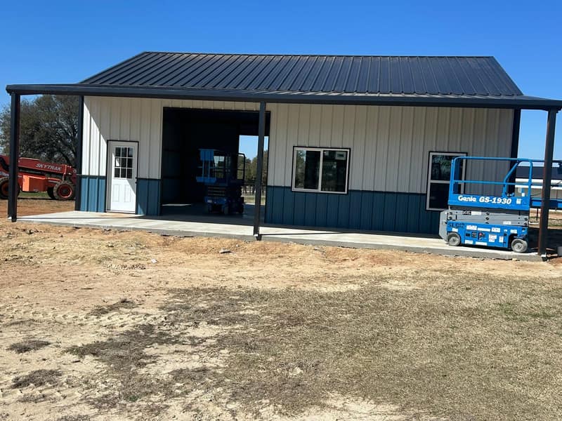 White metal shop with dark blue wainscot, black roof and a covered front porch, scissor lift alongside