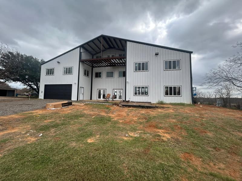 Two-story white metal barndominium with black trim, a second-floor balcony and a garage bay under a stormy sky